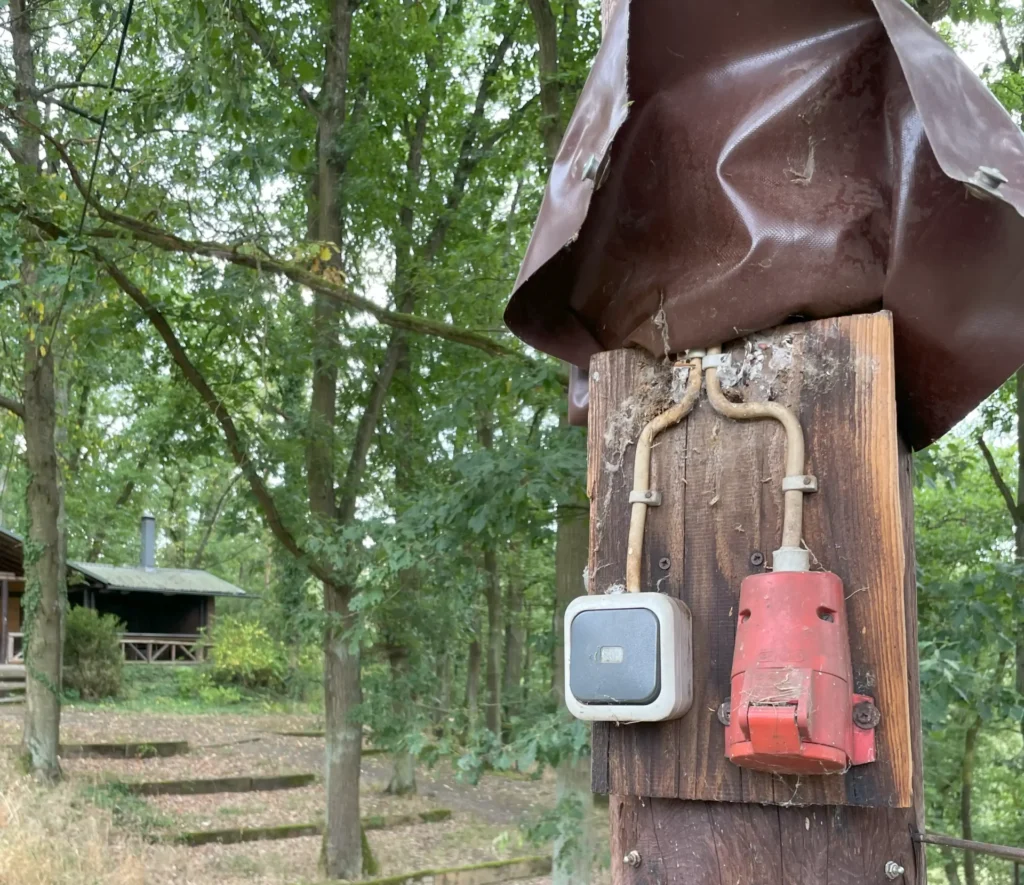 Im Vordergrund ein hölzerner Mast mit einem Schalter und einem roten Drehstrom-Stecker, im Hintergrund Wald und die Schutzhütte