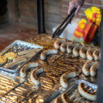 Würstchen auf einem Grill, eine Grillzange ragt in das Bild